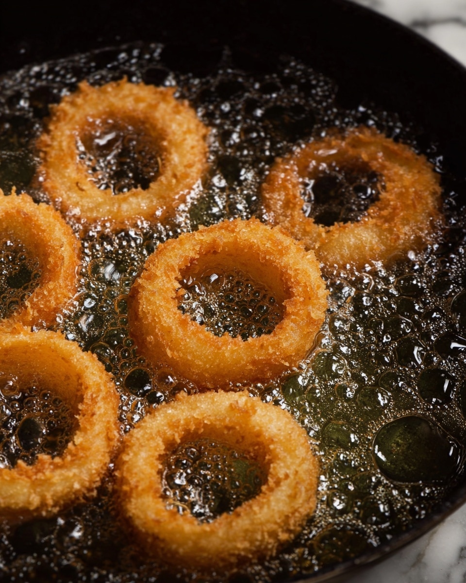 A close-up of a single golden brown, crispy fried onion ring being held by a woman's hand, half dipped in a light orange creamy sauce that drips slowly; the background shows a white marbled surface with out-of-focus kitchen items, and to the right sits a whole light brown onion with skin intact. Photo taken with an iphone --ar 4:5 --v 7