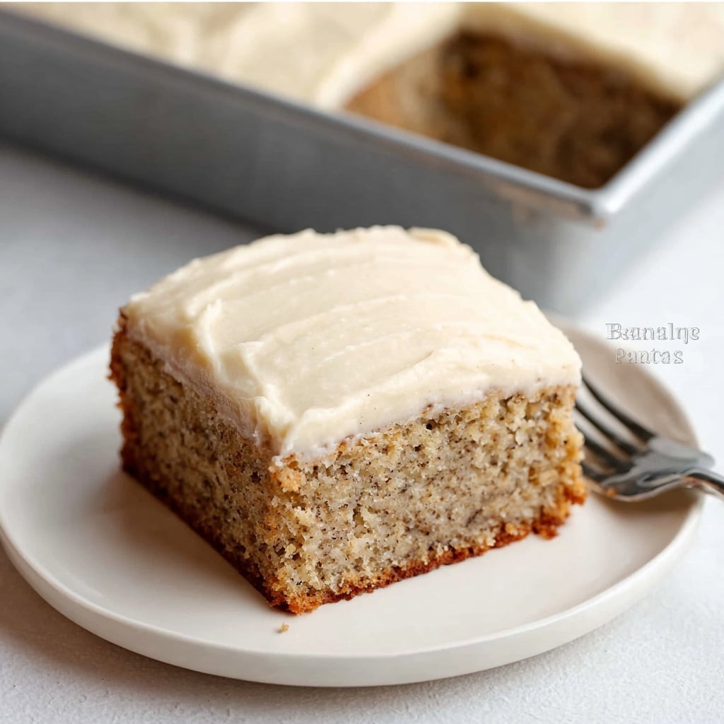 A single square slice of banana cake sits on a white plate, showcasing two distinct layers: the bottom layer is a thick, moist cake with a slightly speckled light brown texture—indicative of mashed banana and spices—while the top layer is a generous, smooth, pale cream-colored frosting evenly spread across the surface. The edges are cleanly cut, revealing the contrast between the denser cake and the fluffy, creamy topping. In the background, the rest of the cake is visible in a metal baking pan, with the same frosting smoothly covering the entire top. photo taken with an iphone --v 7.0