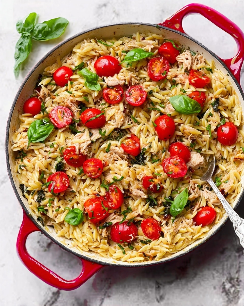 A close-up view of a black pan filled with a dish of grilled chicken pieces, each marked with dark grill lines and topped with green pesto sauce. The chicken sits on a bed of yellowish orzo pasta mixed with pesto, giving it a slightly green color. Scattered bright red cherry tomatoes and fresh green basil leaves add pops of color across the dish. The pan is shown on a white marbled surface. Photo taken with an iphone --ar 4:5 --v 7
