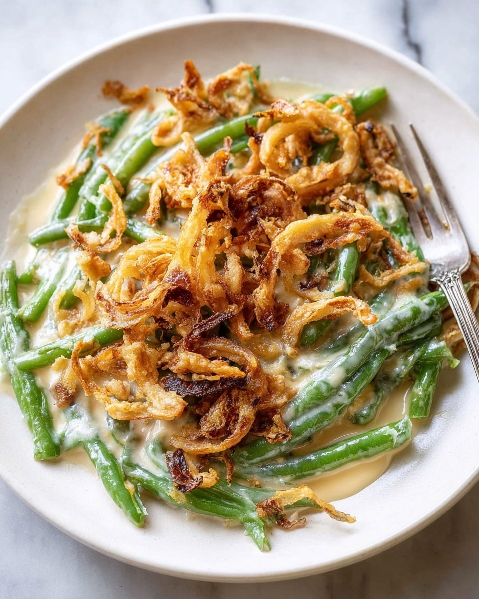 A white round plate holds a green bean casserole with three visible layers. The bottom layer is fresh green beans, bright green and slightly shiny, arranged in various directions. Above the green beans is a creamy, beige cheese sauce covering them unevenly with a smooth and slightly thick texture. The top layer is a generous pile of crispy, golden brown fried onions, curly and uneven with some pieces darker than others, creating a crunchy texture. A metal fork rests on the right edge of the plate. The dish is set on a white marbled surface. Photo taken with an iphone --ar 4:5 --v 7