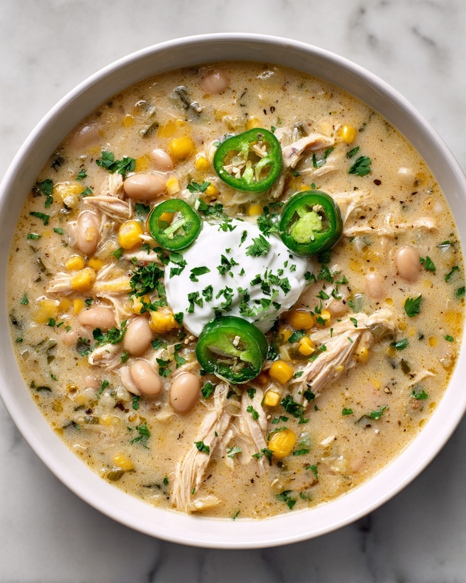 A white bowl is filled with creamy soup with visible textures of shredded chicken, yellow corn kernels, and light brown beans scattered on top. The soup base is light beige with green herbs mixed throughout. There are slices of green jalapeño pepper and chopped green herbs sprinkled all over. In the center, there is a dollop of white sour cream garnished with more green herbs. The bowl rests on a white marbled surface photo taken with an iphone --ar 4:5 --v 7