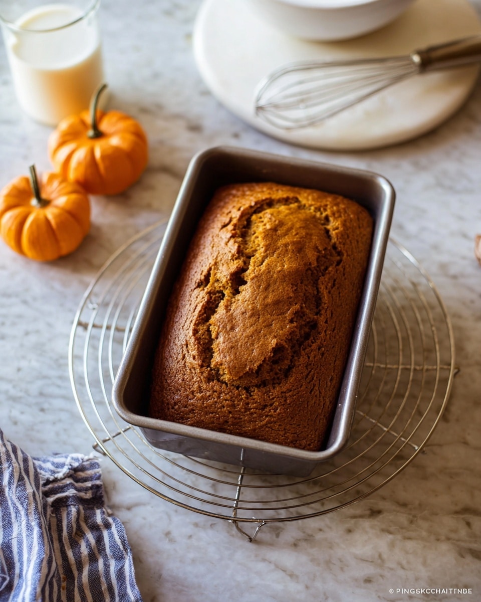 A warm loaf of pumpkin bread rests in a metal baking pan on a round wire cooling rack. The bread has a cracked, golden-brown top with a slightly rough texture and a moist appearance. Around the bread, there are small orange pumpkins, a white marbled surface underneath, a white bowl with a whisk resting on another white marbled round board, and a small glass of milk to the side. A blue and white striped cloth is partially visible in the bottom left corner. The scene has a soft, natural light that highlights the bread's warmth and texture. photo taken with an iphone --ar 4:5 --v 7