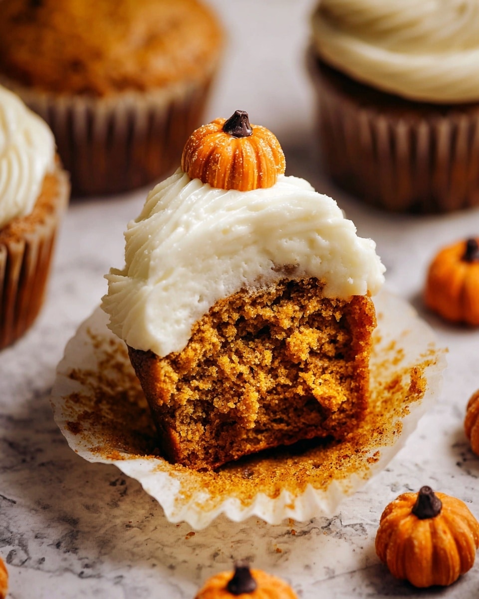 The image shows a partially eaten pumpkin-flavored cupcake sitting in a white paper liner, placed on a white marbled textured surface. The cupcake has two main layers: the bottom layer is moist, crumbly, and orange-brown with a spongy texture, while the top layer is thick, smooth, creamy white frosting swirled in a wave-like shape. On top of the frosting sits a small, round, pumpkin-shaped decoration with an orange, slightly rough texture and a small dark chocolate point on top. Around the cupcake, there are several more small pumpkin-shaped decorations scattered, each with a ridged texture and dark center, adding a festive autumn feel. photo taken with an iphone --ar 4:5 --v 7