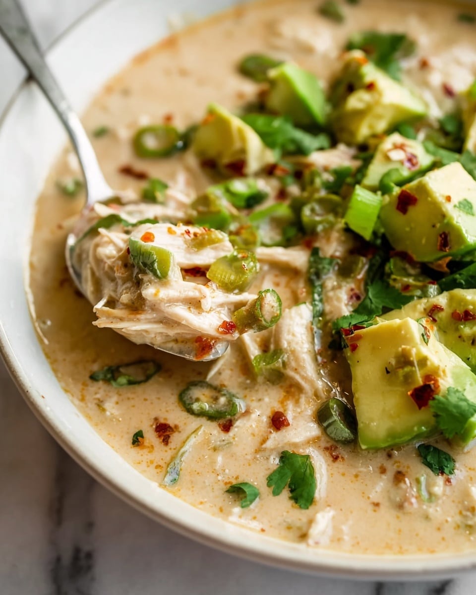 The image shows a close-up of a creamy soup in a white bowl set on a white marbled surface. The soup has a thick, beige creamy base with shredded white chicken mixed in. On top, there are bright green cubes of avocado, chopped green onions, small cilantro leaves, and bits of red pepper flakes adding color and texture. A silver spoon is scooping up the soup, holding a creamy mix of chicken and sauce with bits of green and red. The overall look is rich, smooth, and fresh with vibrant green and warm beige tones. photo taken with an iphone --ar 4:5 --v 7