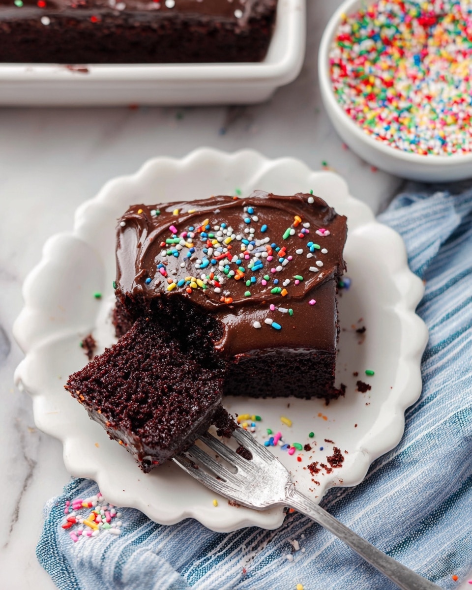 A square slice of chocolate cake with one thick, moist, and dark brown layer topped with a smooth, shiny layer of dark chocolate frosting spread evenly. Colorful round sprinkles in red, white, green, yellow, blue, pink, and orange are sprinkled over the frosting. A piece of cake has been cut and is held on a fork showing the crumbly and soft texture inside. The cake rests on a white plate with scalloped edges, set on a white marbled surface with a blue and white striped cloth nearby. A bowl filled with more colorful round sprinkles sits in the background. photo taken with an iphone --ar 4:5 --v 7
