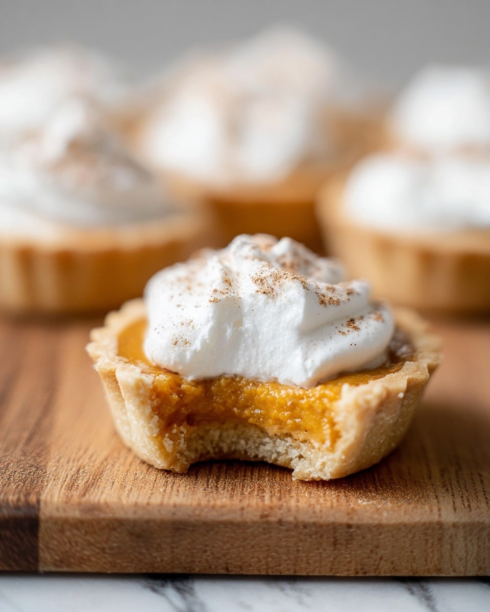 A close-up of a small tart held by a woman's hand, showing three main layers: a light brown, crumbly crust forming the base and edges; a smooth, shiny orange filling covering the crust; and a fluffy white whipped cream dollop on top, sprinkled with a light dusting of brown cinnamon powder. The background is softly blurred with similar tarts in soft focus on a white marbled surface. photo taken with an iphone --ar 4:5 --v 7