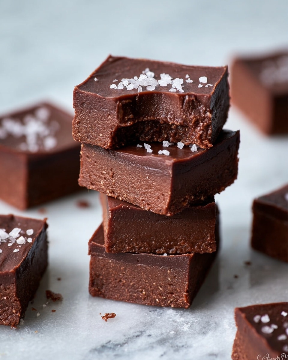 The image shows a stack of four thick chocolate fudge squares on a white marbled surface. Each square has two layers: a dense, rich dark brown bottom layer with a slightly crumbly texture, and a smoother, glossy lighter brown top layer. The top square has a bite taken out of one corner, revealing the creamy inside of the top layer and the firm bottom layer beneath. Small coarse salt crystals are sprinkled on the top layer of the top square, adding a shiny texture. Four more fudge squares are scattered around the stack in the background, slightly blurred. photo taken with an iphone --ar 4:5 --v 7