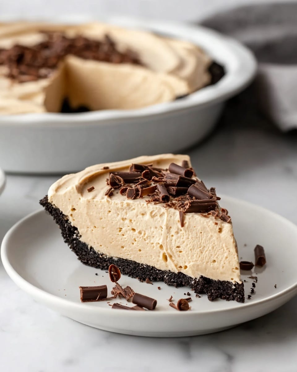 A slice of pie sits on a white plate on a white marbled surface, showing three layers: a crumbly dark brown cookie crust at the bottom, a thick smooth light beige creamy filling in the middle with an airy texture, and a slightly swirled creamy beige top layer. The top is decorated with several dark chocolate curls scattered across the slice and around the plate. In the background, the rest of the pie in a white dish is visible, with one slice missing. photo taken with an iphone --ar 4:5 --v 7