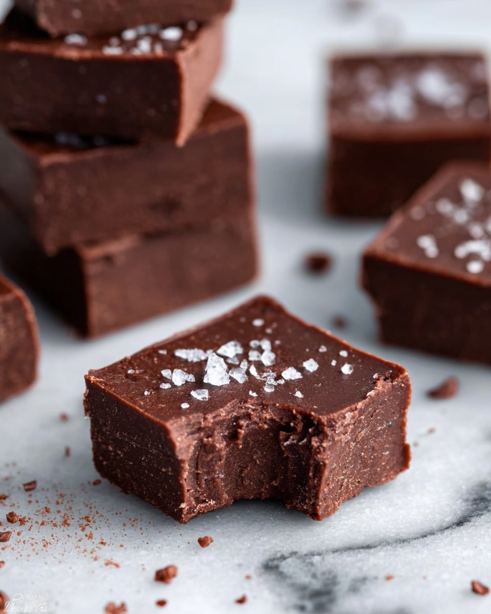 A close-up image shows several square-shaped chocolate fudge pieces on a white marbled surface, with one piece in the front having a bite taken out of it. The fudge is dark brown and smooth with a glossy top layer, sprinkled with coarse salt crystals. Behind it, there is a stack of multiple fudge squares, showing a thick, dense texture with slightly rough edges. Around the pieces are small scattered cocoa powder bits and salt crystals. The photo taken with an iphone --ar 4:5 --v 7