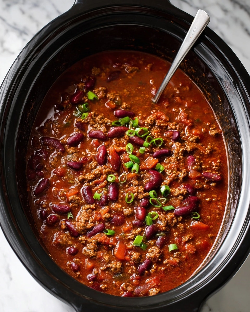 A black slow cooker filled with a thick, rich chili showing one layer of chunky texture with visible dark red kidney beans scattered evenly throughout, mixed with small browned ground meat pieces and soft translucent onion pieces, all swimming in a deep reddish-brown tomato-based sauce with hints of green chopped scallions sprinkled on top. A spoon is partially submerged in the chili on the left side. The cooker is set on a white marbled surface. photo taken with an iphone --ar 4:5 --v 7