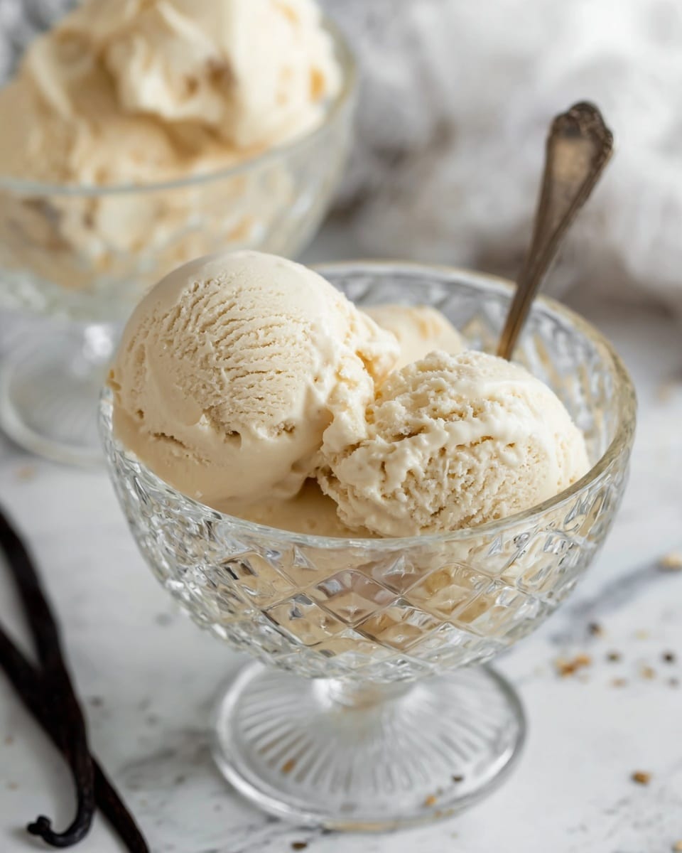 Two scoops of creamy, light beige ice cream with a smooth yet slightly textured surface are served in a clear glass dessert bowl with a diamond-cut pattern. The scoops sit close together, filling the bowl, with a silver spoon partially inserted into the ice cream. In the background, there is a second similar bowl filled with more ice cream, slightly blurred. The setting is on a white marbled surface with a vanilla bean pod and some scattered vanilla seeds nearby. photo taken with an iphone --ar 4:5 --v 7
