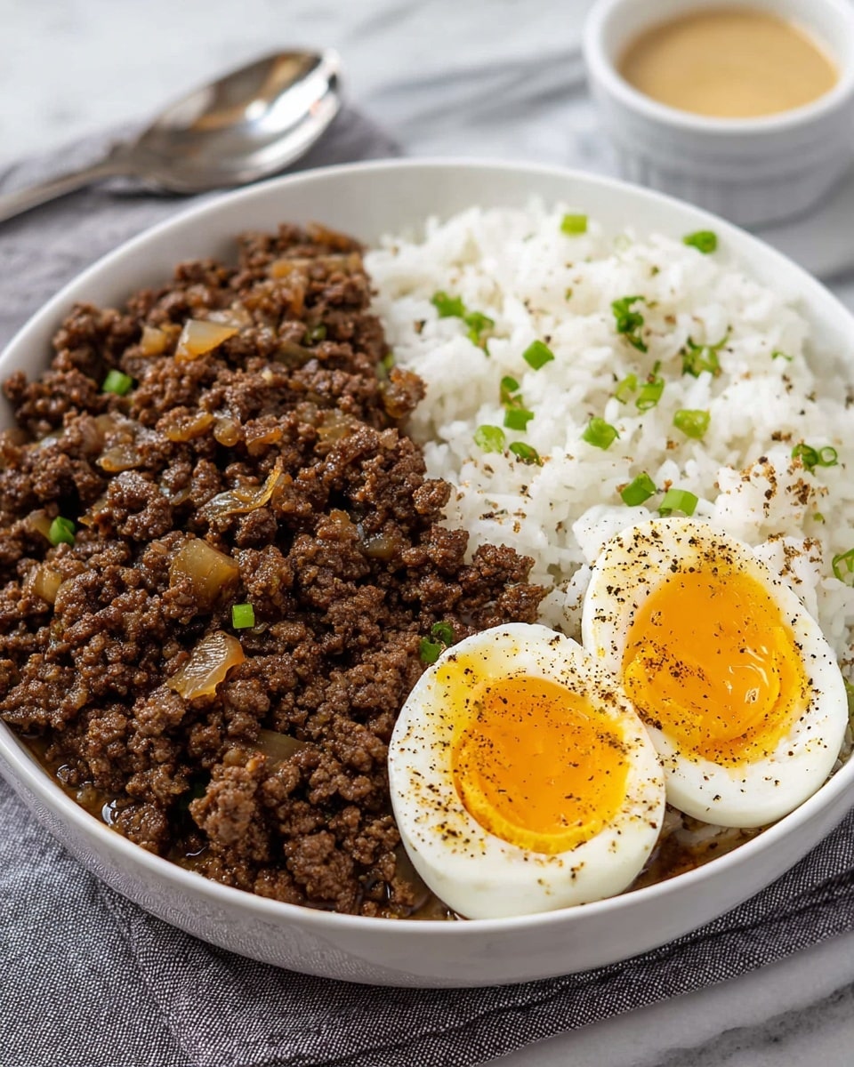 A white bowl holds three main parts: on the left, a large portion of cooked ground beef mixed with small bits of cooked onion, dark brown with a slightly glossy texture; to the right, a fluffy mound of white rice with scattered green onion pieces; in front, two halves of a soft-boiled egg with firm white edges and bright, runny golden yolks sprinkled with black pepper. The bowl sits on a folded gray cloth on a white marbled surface, with part of a silver spoon visible at the right edge and a small container of light-colored sauce blurred in the background. photo taken with an iphone --ar 4:5 --v 7