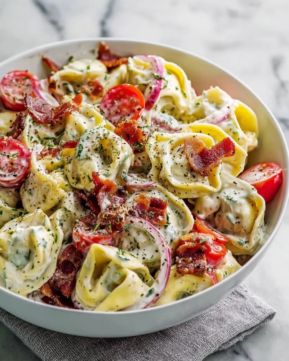 A white bowl filled with creamy tortellini pasta salad, showing about four layers of folded pasta in pale yellow color mixed with bright red cherry tomato halves and thin red onion rings. Scattered on top are crispy, dark reddish-brown bacon pieces and slices of yellow-green pepper rings. The salad is coated in a thick white creamy dressing with green herb specks and a light sprinkle of black pepper. The bowl sits on a light gray cloth against a white marbled surface. photo taken with an iphone --ar 4:5 --v 7