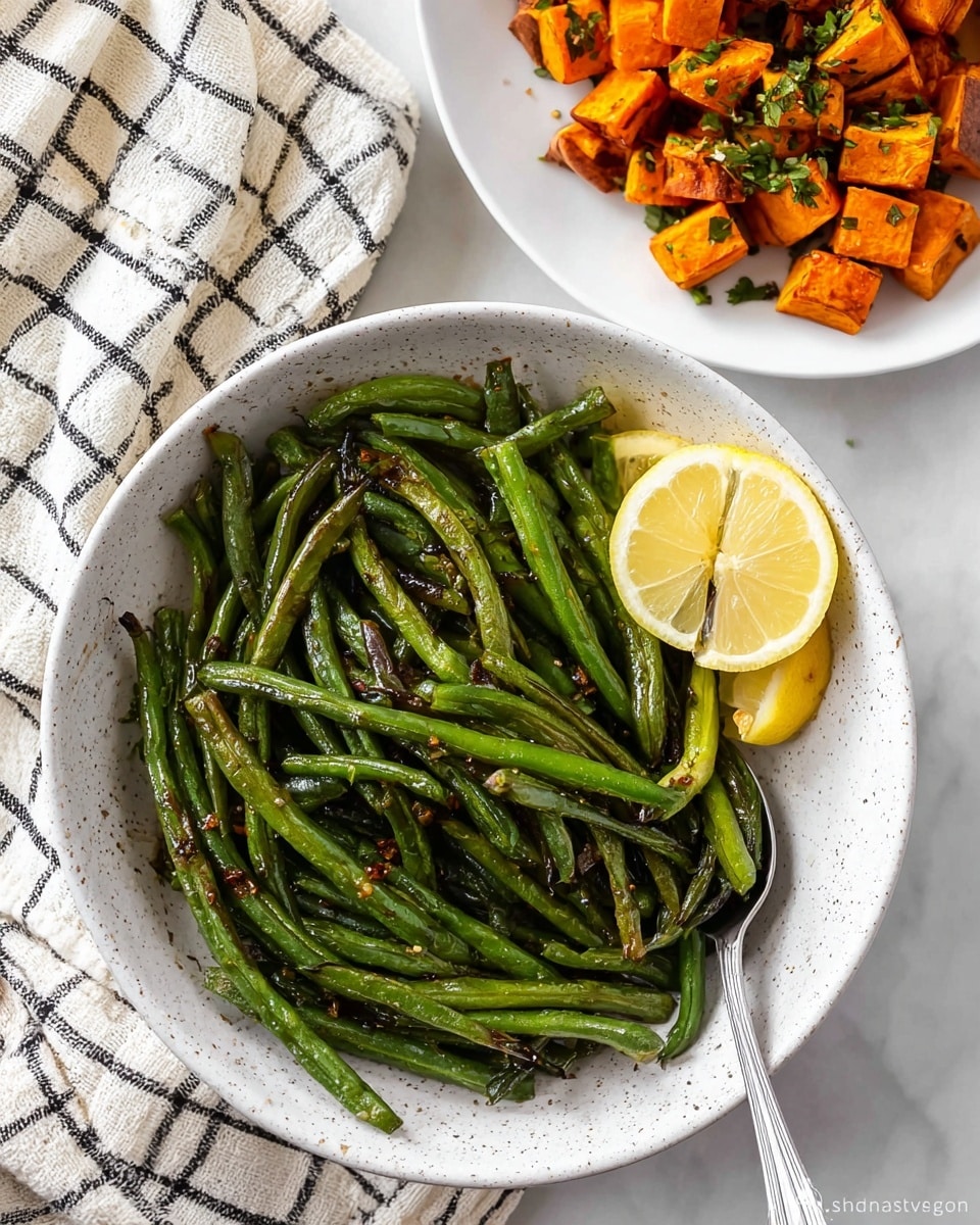A white speckled bowl filled with cooked green beans forms the main part of the image. The green beans are shiny and slightly charred, scattered in a natural pile, showing different lengths and thicknesses with small bits of seasoning on top. Two lemon wedges, pale yellow with a soft texture, rest on the right edge of the green beans. A silver serving spoon peeks from underneath the green beans at the bottom of the bowl. In the upper right corner, a white plate with roasted, cubed sweet potatoes is partially visible, the orange cubes showing clear browned spots with small green herb pieces on top. A cream-colored cloth with black checked lines lies crumpled on the upper left of the scene. All items rest on a white marbled surface. Photo taken with an iphone --ar 4:5 --v 7