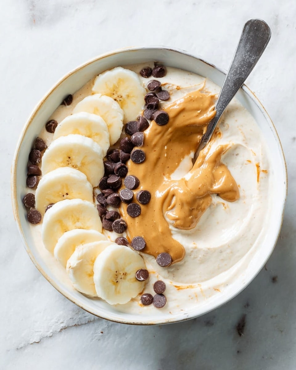 A white bowl filled with a creamy, off-white base layered smoothly on the bottom. On top, there are two rows of banana slices placed on one side of the bowl, showing a light yellow color with a soft texture. A thick, golden-brown peanut butter layer is spread in two parts, one small patch near the center and one drizzle over the banana slices. Scattered throughout the bowl are small, round dark brown chocolate chips adding texture and contrast. A spoon rests inside the bowl, pushing into the creamy layer, and the bowl sits on a white marbled surface. Photo taken with an iphone --ar 4:5 --v 7