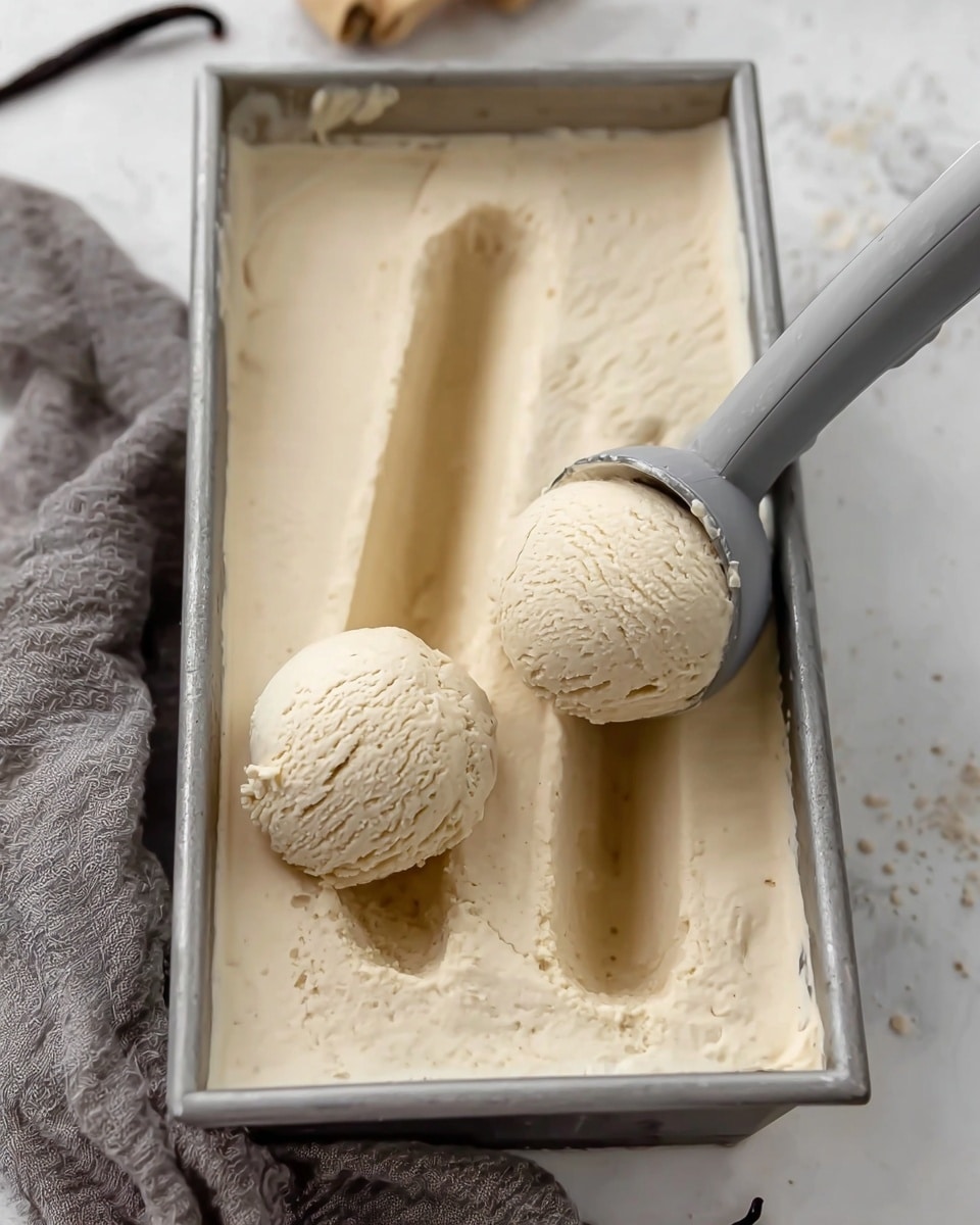 A rectangular metal container filled with smooth, creamy beige ice cream, showing clear soft texture and tiny vanilla bean specks throughout. Two rounded scoops of the ice cream are being lifted from the container with a gray ice cream scoop, leaving two deep, curved grooves on the surface of the ice cream. The metal container sits on a white marbled texture surface with a gray cloth partially visible in the background. photo taken with an iphone --ar 4:5 --v 7