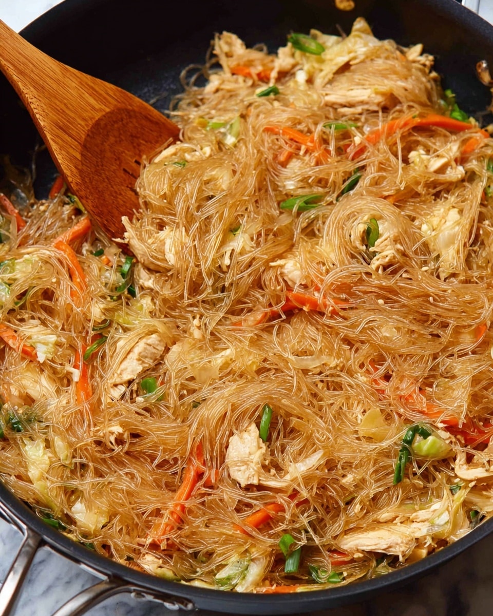 A close-up of a cooked dish inside a black pan shows thin, shiny glass noodles mixed with various vegetables and pieces of chicken. The noodles are light brown and tangled, making the main layer. Interspersed throughout are bright orange carrot strips, white onion slices, green onion pieces, and light green cabbage, adding color and texture. The chicken pieces are white and small, sitting near the bottom and middle layers, blending with the vegetables. A wooden spoon rests on the left side, partially scooping the noodles. The overall look is shiny and slightly oily, indicating the dish is freshly cooked. The background surface is a white marbled texture. photo taken with an iphone --ar 4:5 --v 7