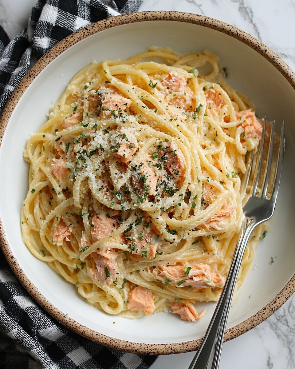 A neat mound of spaghetti pasta sits in the center of a large white plate, with a creamy light yellow sauce coating the noodles. Thin slices of pink smoked salmon are layered on top and mixed slightly with the pasta. Bright green chopped chives are sprinkled over the salmon and pasta, adding a fresh touch. Small black pepper flakes are scattered around the plate and on the noodles. A shiny silver fork and spoon rest together on the right side edge of the plate. The plate is set on a white marbled surface next to a folded light gray cloth napkin. photo taken with an iphone --ar 4:5 --v 7