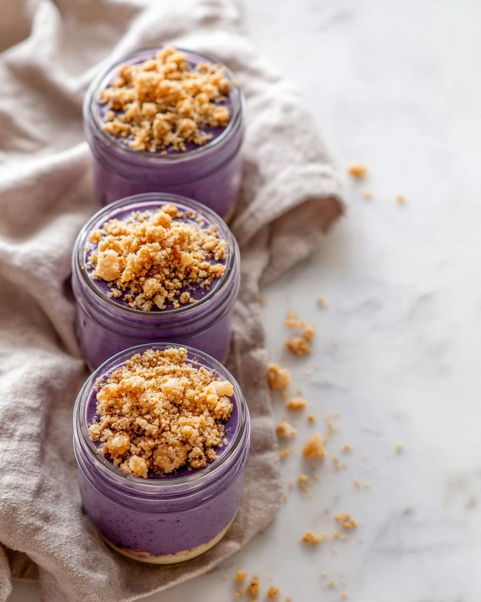 Three small glass jars are filled with two layers: a bottom layer of thick purple mousse and a top layer of crumbly golden brown streusel. The jars sit vertically aligned on a beige cloth over a white marbled surface, with scattered crumbs around. The image is bright and clear, showing the texture contrast between the smooth mousse and the crunchy topping. photo taken with an iphone --ar 4:5 --v 7
