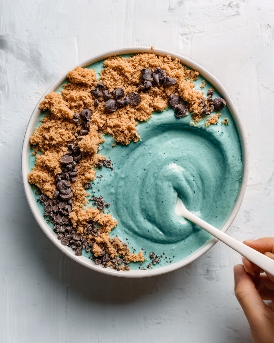 A white bowl filled with three distinct layers is placed on a white marbled surface. The bottom layer is a smooth, thick teal cream or smoothie that fills most of the bowl. On top of the right side of this teal base, there is a rough, crumbly layer of light brown cookie dough or crumble, spreading partially and unevenly over the teal surface. Scattered over the brown layer and partly extending into the teal area are small, dark brown chocolate chips or chunks that add texture and contrast. A woman's hand holds a white spoon, dipping into the teal layer near the center of the bowl. Photo taken with an iphone --ar 4:5 --v 7