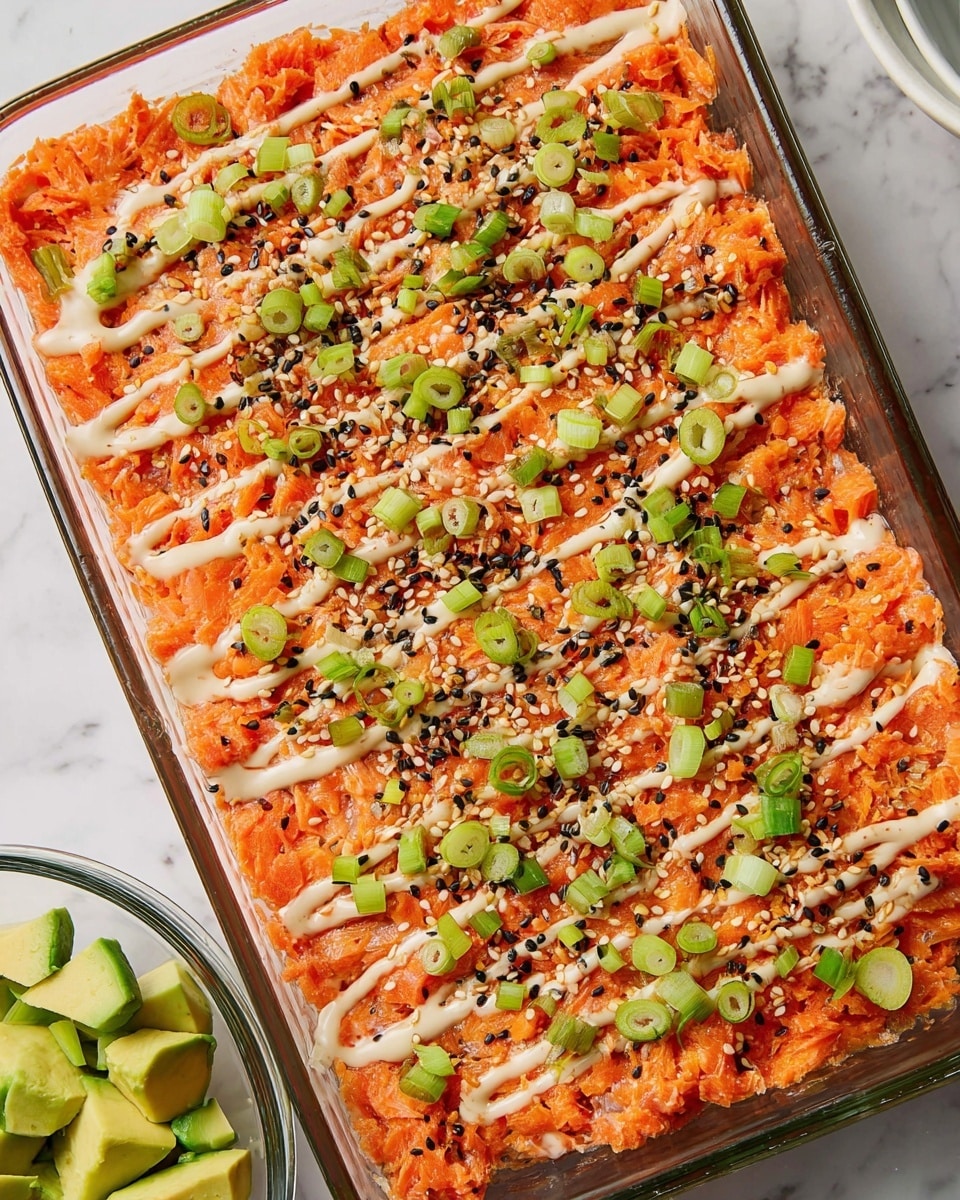 A rectangular glass dish filled with an evenly spread, bright orange layer of shredded or mashed cooked salmon, textured with small chunks and flakes. Over the top, there are thin, zigzagging lines of light cream-colored sauce. Scattered all over the surface are small, round slices of green onions and a mix of black and white sesame seeds, adding contrast and texture. To the side, there is a clear bowl with diced green avocado pieces resting on a white marbled surface. The overall presentation is neat, colorful, and fresh-looking. Photo taken with an iphone --ar 4:5 --v 7
