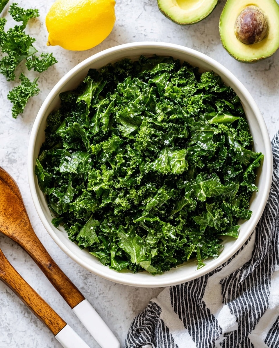 A large white bowl filled with finely chopped fresh kale, showing deep green curly leaves with a slightly shiny texture from light dressing. Around the bowl, there is a whole yellow lemon on the white marbled surface, two wooden salad utensils with white handles placed diagonally, a striped cloth napkin in black and white, and two halves of a ripe avocado with dark green skin and bright green flesh with the seed in one half. Photo taken with an iphone --ar 4:5 --v 7
