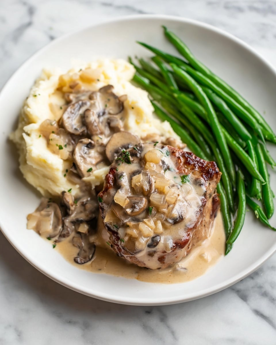 A white plate holds a meal with three main parts arranged side by side: on the left, a generous scoop of creamy mashed potatoes covered in a pale beige mushroom sauce with visible sliced mushrooms; in the center, a thick, round piece of browned meat topped with the same creamy mushroom sauce containing diced onions and herbs; on the right, a neat row of bright green steamed green beans. The background is a white marbled surface. Photo taken with an iphone --ar 4:5 --v 7