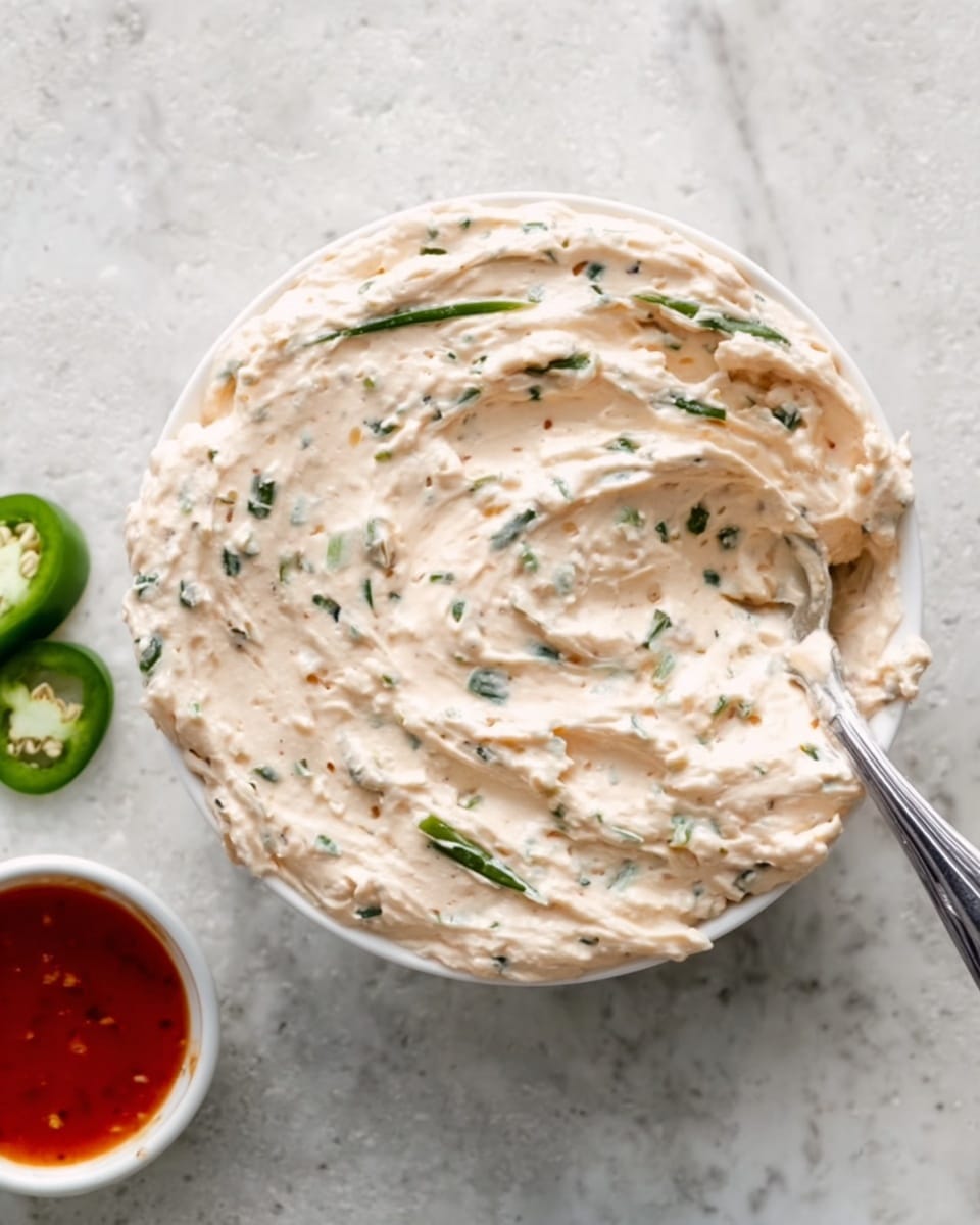 A white bowl filled with a creamy light beige spread that has small green herb pieces mixed in, creating a speckled pattern throughout. A silver spoon is partially scooping some of the spread from the right side of the bowl. To the left, part of a small white bowl with a red sauce and two green jalapeño slices are visible. Everything is set on a white marbled textured surface. photo taken with an iphone --ar 4:5 --v 7