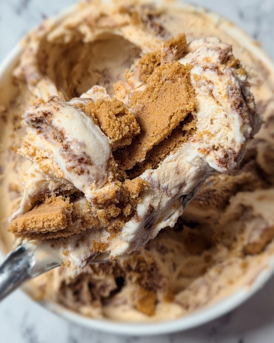 The image shows a close-up of a creamy dessert with visible layers of soft, light brown cookie pieces mixed into a thick, beige cream with brown speckles. The texture appears gooey and slightly chunky, with broken cookie bits layered unevenly within the creamy mixture. The dessert is held on a silver spoon lifting a portion from a white bowl resting on a white marbled surface. The overall look is rich and dense, with contrasting rough cookie crumbs and smooth cream blending closely together. photo taken with an iphone --ar 4:5 --v 7