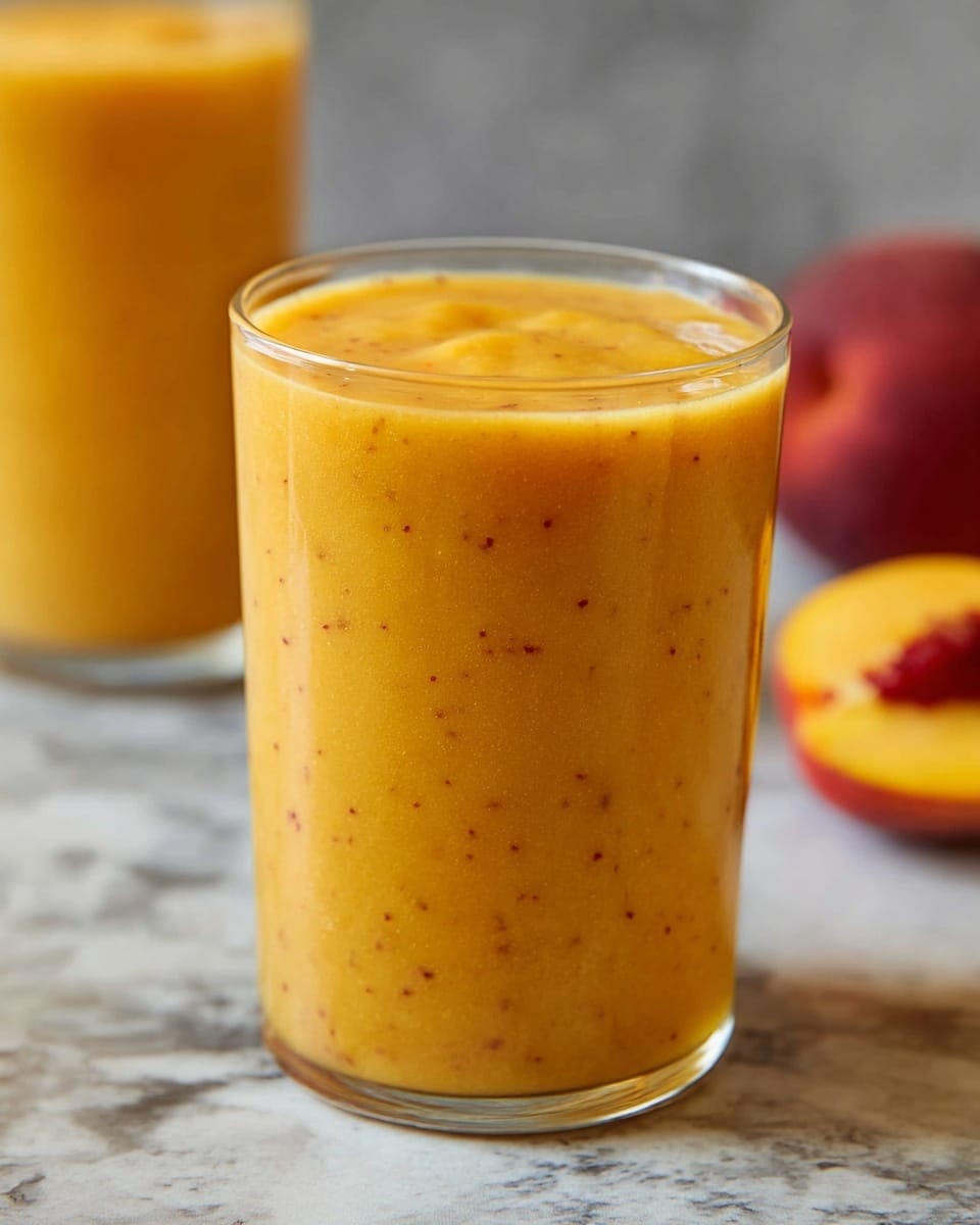 A close-up image of a clear glass filled with a thick, creamy smoothie that has a bright orange color with small dark specks evenly spread throughout. The smoothie fills the glass almost to the top with a smooth, slightly shiny surface. Behind the glass, there is another taller glass with the same smoothie, blurred out, and in the background, a halved peach with a yellow flesh and reddish skin rests on a white marbled texture. photo taken with an iphone --ar 4:5 --v 7