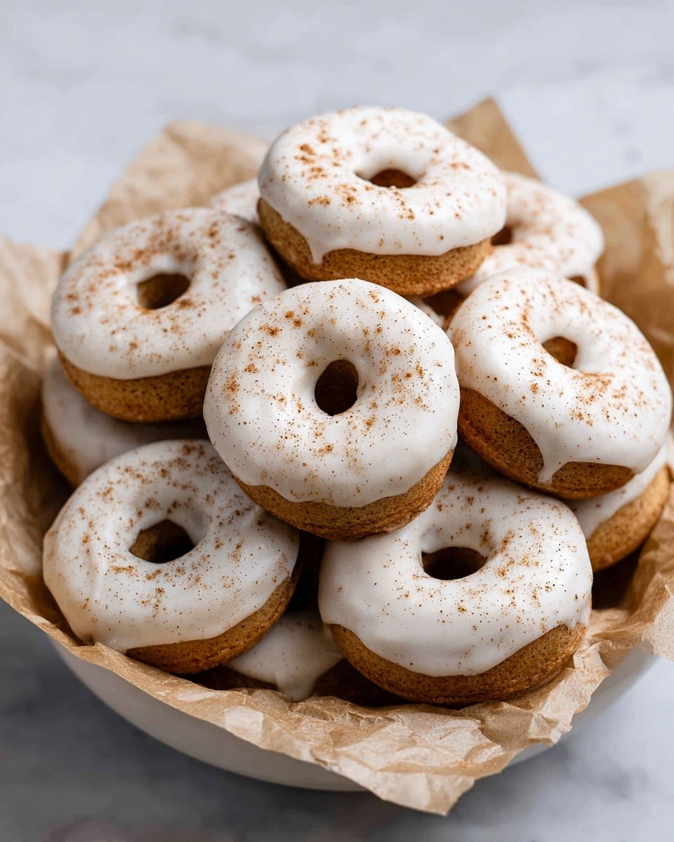 A group of round donuts with a light brown base is piled in a white bowl lined with crinkled light brown parchment paper. Each donut is topped with a thick, smooth white icing layer that covers the top surface and drips slightly over the edges. The icing is sprinkled with a fine dusting of brown spice, creating a soft speckled effect. The donuts are stacked in multiple layers, some overlapping, filling the bowl evenly. The entire scene sits on a white marbled surface. photo taken with an iphone --ar 4:5 --v 7