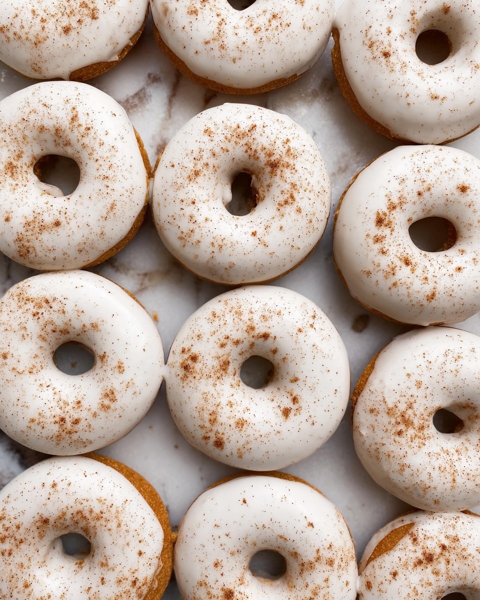 The image shows a group of a dozen round donuts with a hole in the middle, all placed close together on a white marbled surface. Each donut has a thick layer of smooth white icing covering the top, with a light sprinkling of fine brown powder, possibly cinnamon, scattered evenly over the icing. The base of the donuts is a warm golden brown, visible around the edges just beneath the white icing. The donuts are arranged in neat rows, filling the frame with a uniform and inviting look. Photo taken with an iphone --ar 4:5 --v 7