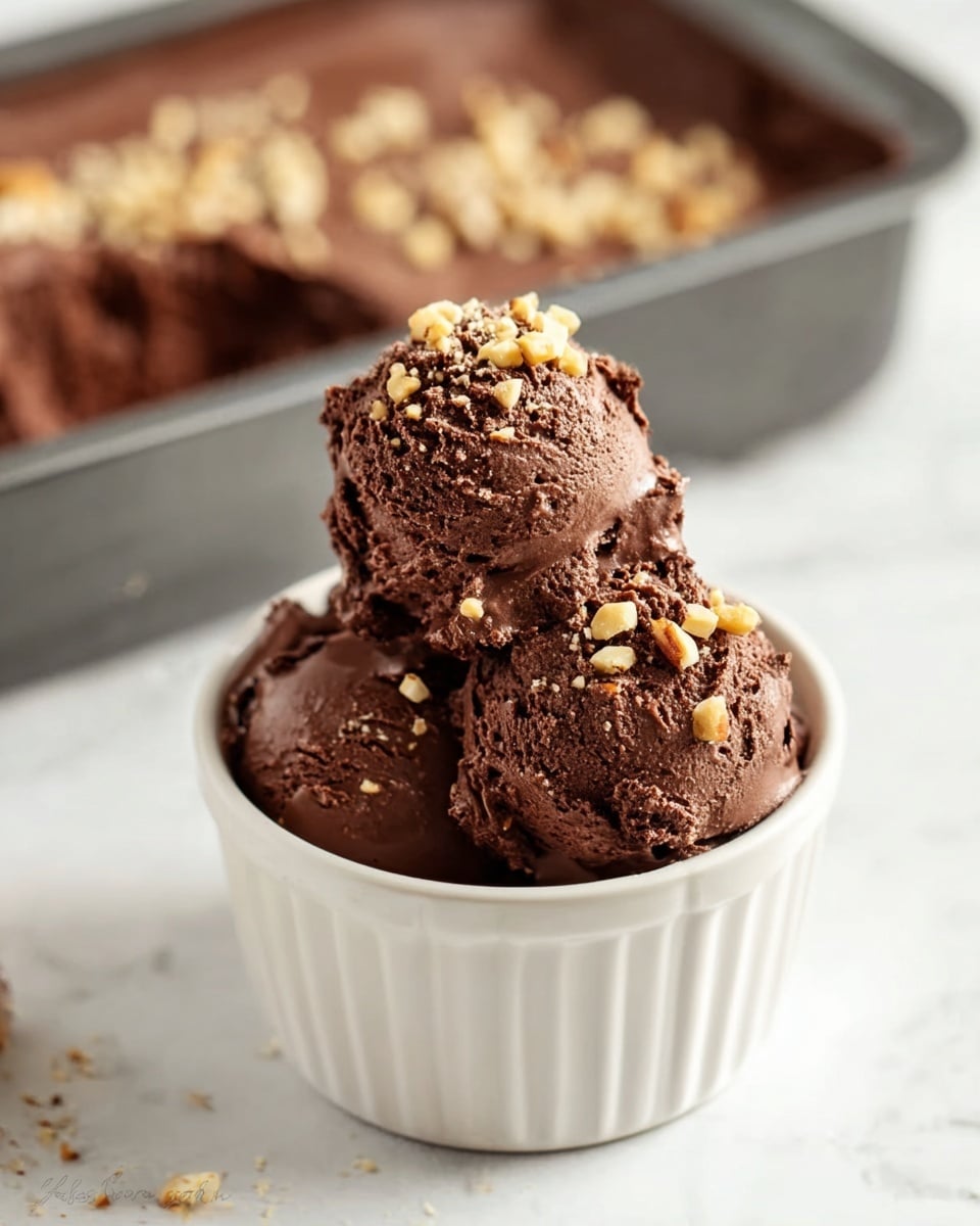 The image shows three scoops of rich, dark brown chocolate ice cream stacked inside a small white ceramic bowl with vertical ridges. The ice cream looks creamy with a slightly rough texture and is topped with small light brown nut pieces scattered over all scoops. In the blurred background, part of a rectangular baking dish filled with more chocolate ice cream sprinkled with nuts is visible. The scene is set on a white marbled surface. photo taken with an iphone --ar 4:5 --v 7