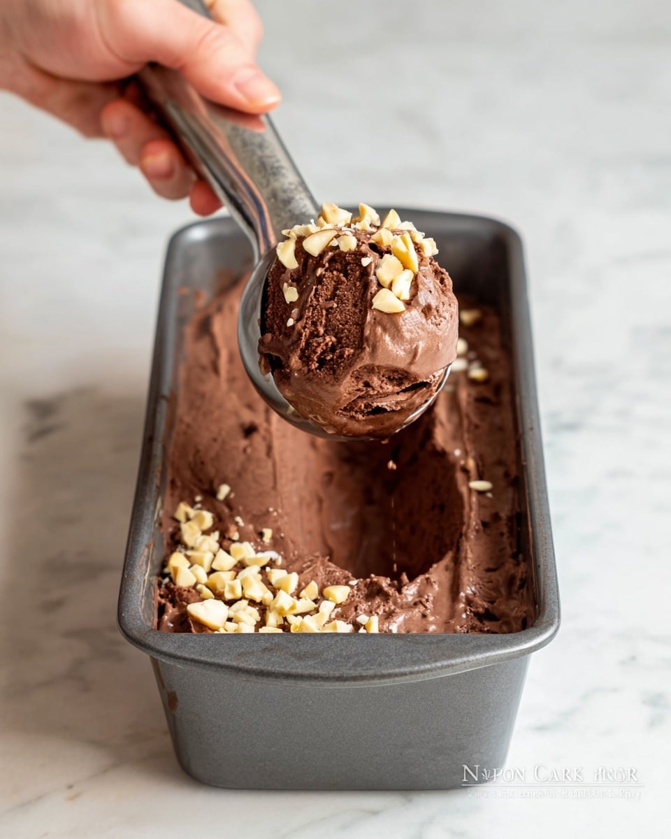A dark brown chocolate ice cream with a creamy and smooth texture is shown being scooped from a gray loaf pan with a metal ice cream scoop. The ice cream is topped with small pieces of chopped nuts, adding a light tan color and crunchy texture on top of the thick chocolate layer. The background is a white marbled surface. A woman's hand is holding the scoop above the pan, lifting a portion of ice cream. The pan is about half empty with some ice cream clinging to the edges. Photo taken with an iphone --ar 4:5 --v 7