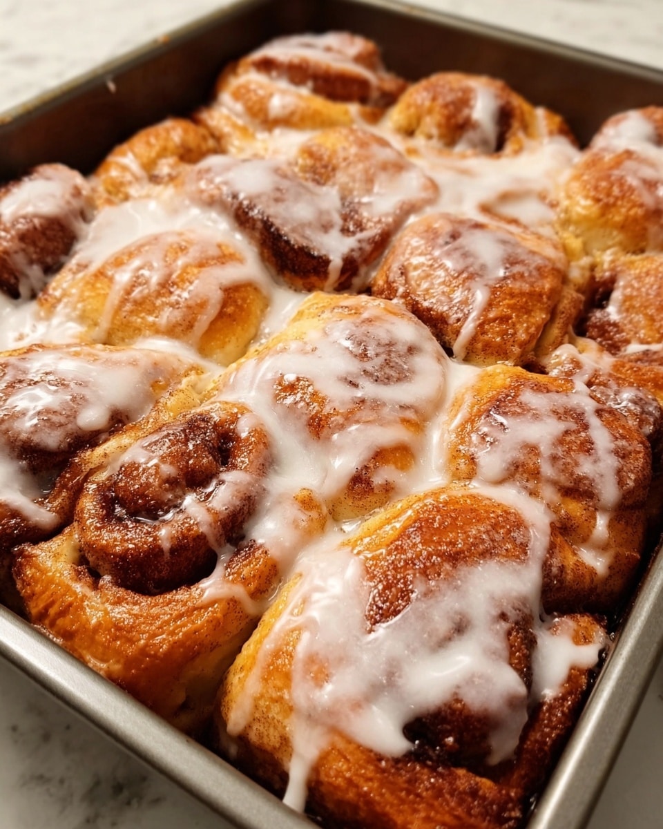 A close-up image of a rectangular baking pan filled with golden brown baked cinnamon rolls. The rolls are tightly packed in one layer, showing a soft texture and a slightly crispy outer crust. A thick layer of white icing is drizzled unevenly over the top, melting and pooling in the gaps between the rolls. The pan sits on a white marbled surface. Photo taken with an iphone --ar 4:5 --v 7