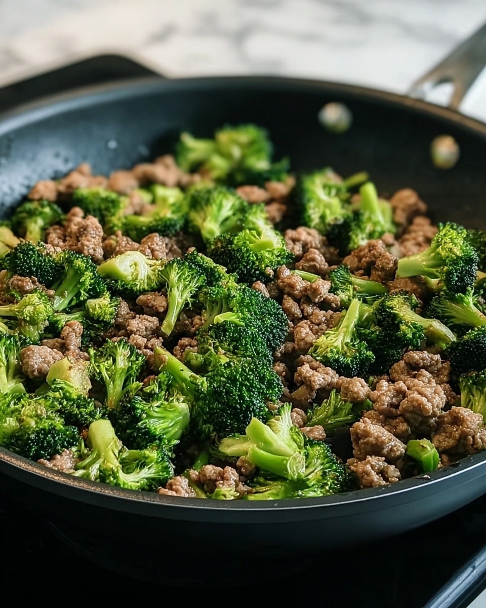 A close-up view of a black frying pan filled with cooked ground meat and bright green broccoli florets mixed evenly throughout. The meat pieces are small, browned with a slightly crumbly texture, while the broccoli pieces maintain their vibrant green color and slightly crisp look. The pan sits on a stove with a blurred white marbled texture background, giving a clean and fresh kitchen feel. Photo taken with an iphone --ar 4:5 --v 7