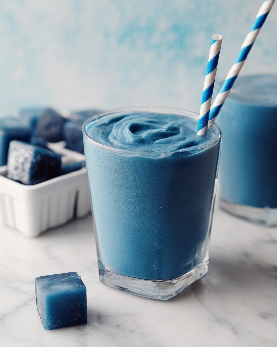 A clear glass filled with a thick, smooth, blue smoothie with a slightly raised swirl on top. A striped blue and white paper straw is inserted on the right side. In the background, there is a white bowl with dark blue ice cubes and a white ice tray with blue ice cubes, all set on a white marbled surface. Photo taken with an iphone --ar 4:5 --v 7