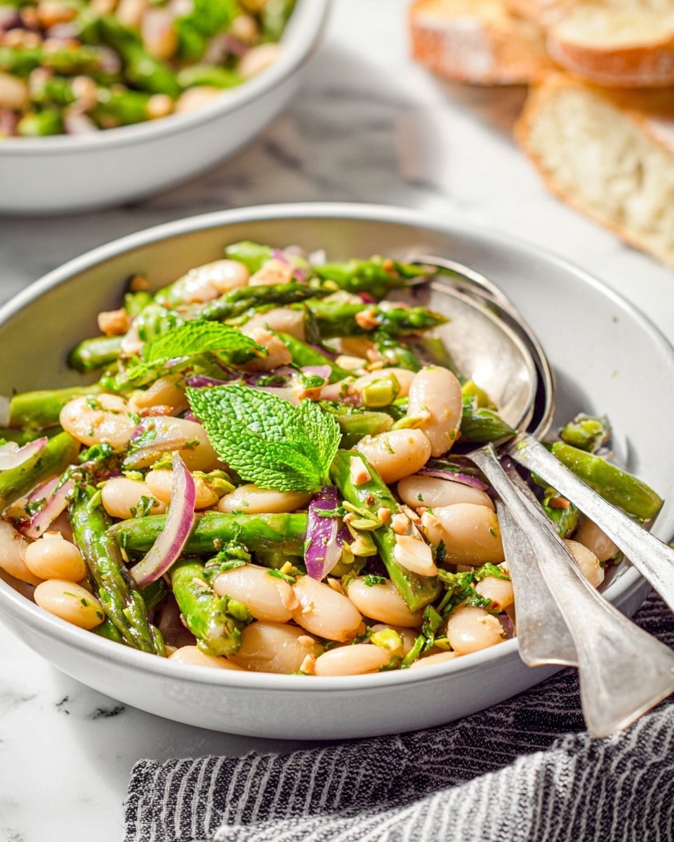 The dish is a vibrant bean and vegetable salad served in a white bowl, filled with three main layers: the bottom layer consists of tender white beans, the middle layer features chopped bright green asparagus pieces mixed with thin slices of translucent red onions, and the top layer is garnished with fresh green mint leaves and small bits of nuts sprinkled over the salad. A silver spoon and a silver fork rest inside the bowl, slightly angled in opposite directions. The bowl is placed on a white marbled surface with a striped cloth partially visible beneath it. In the background, there is a blurred view of another white bowl with a similar salad and a piece of crusty bread. Photo taken with an iphone --ar 4:5 --v 7
